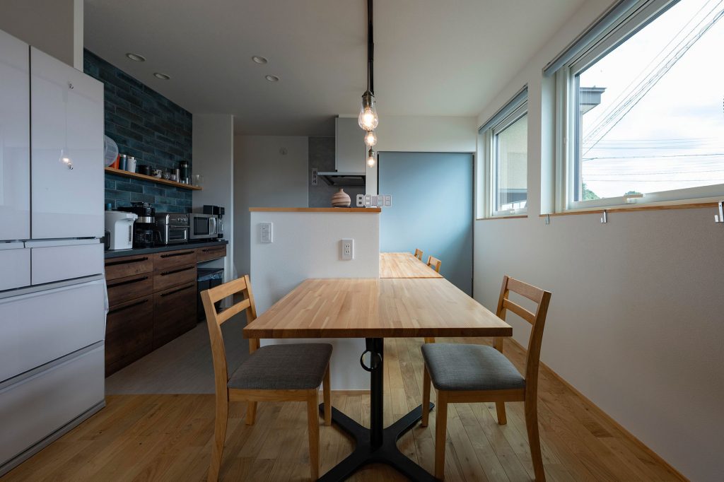 クリ材の無垢床に白壁でナチュラルにまとめられたダイニング・キッチン。背面収納の風合いが美しいタイル壁が空間を彩る。右奥の水色は収納を隠すロールスクリーン
